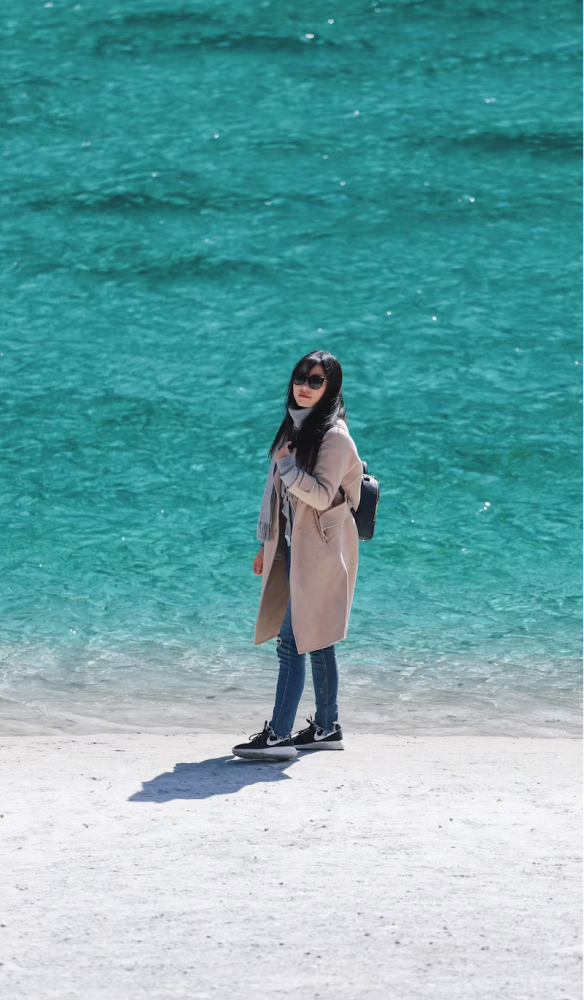 A woman in a long jacket and jeans standing on a beach, right up against the shoreline, showing off her sneakers