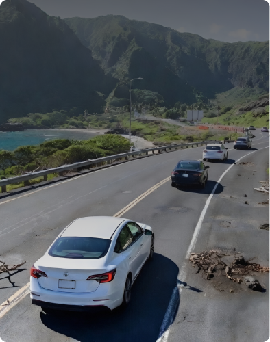 Cars driving along a coastal road with mountains in the background.