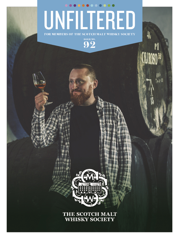 A man holding a glass of whisky, standing near barrels.
