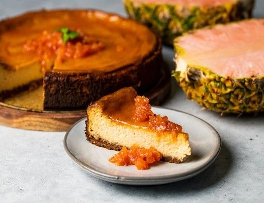 Cheesecake slice with topping, whole cheesecake, and pineapples in the background.