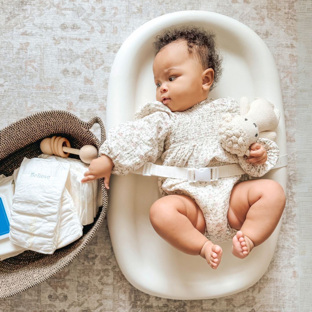 Baby lying in a bassinet, holding a stuffed animal next to a diaper basket.