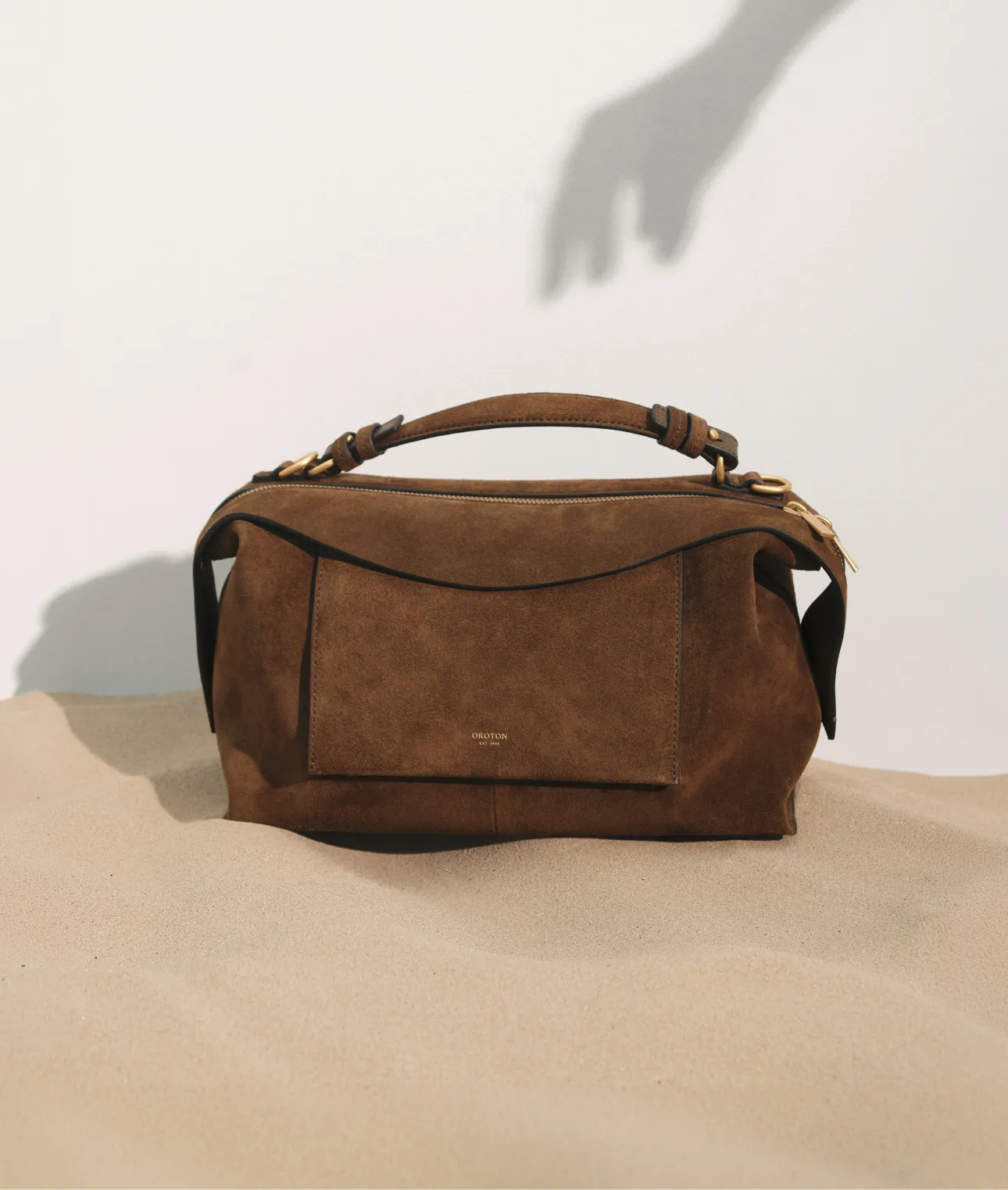 Brown suede handbag on a sandy surface with a hand shadow.