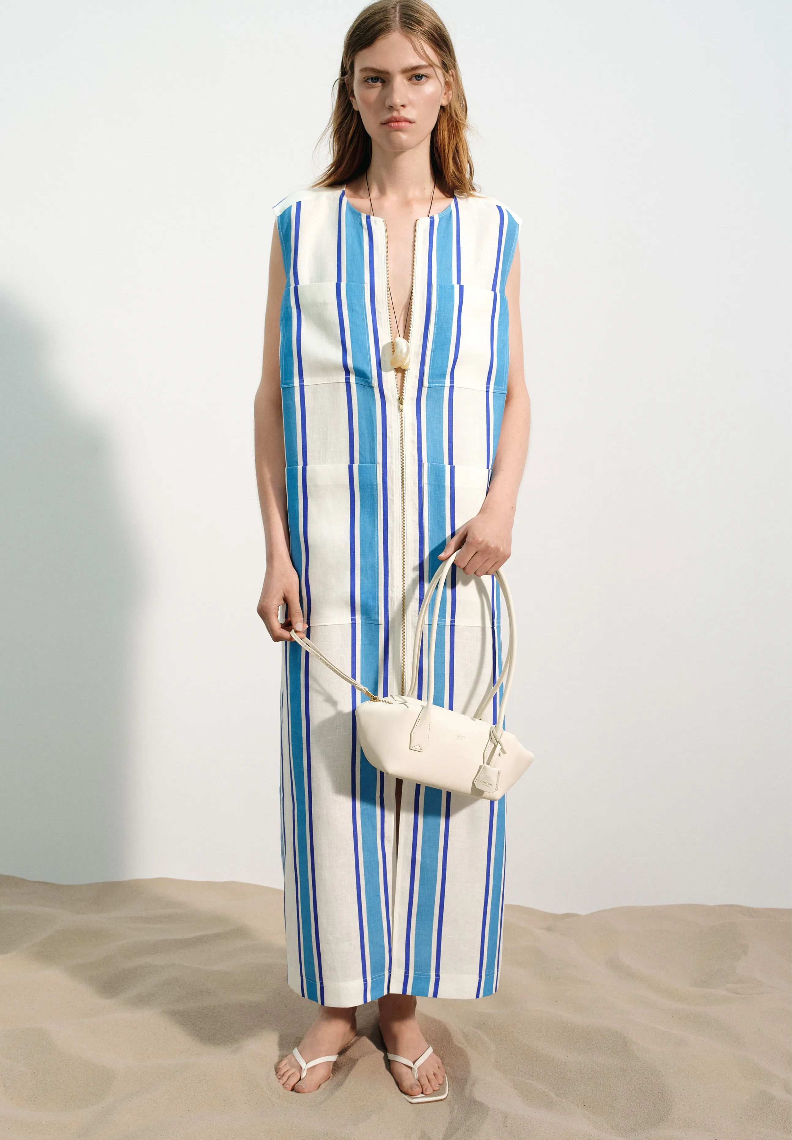 Model wearing a striped dress and holding a handbag on a sandy background.