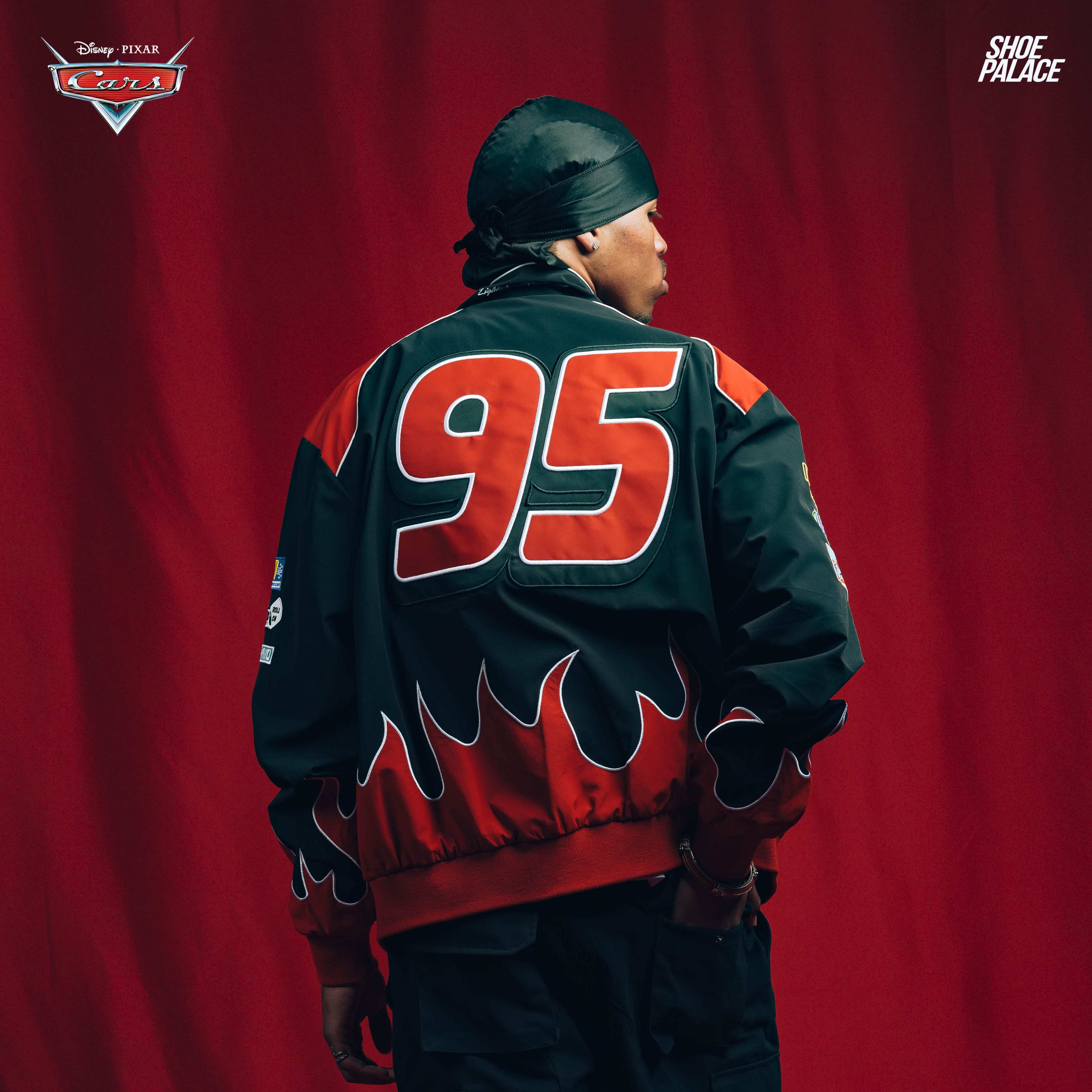 Person in a black and red jacket with flames, facing away against a red backdrop.