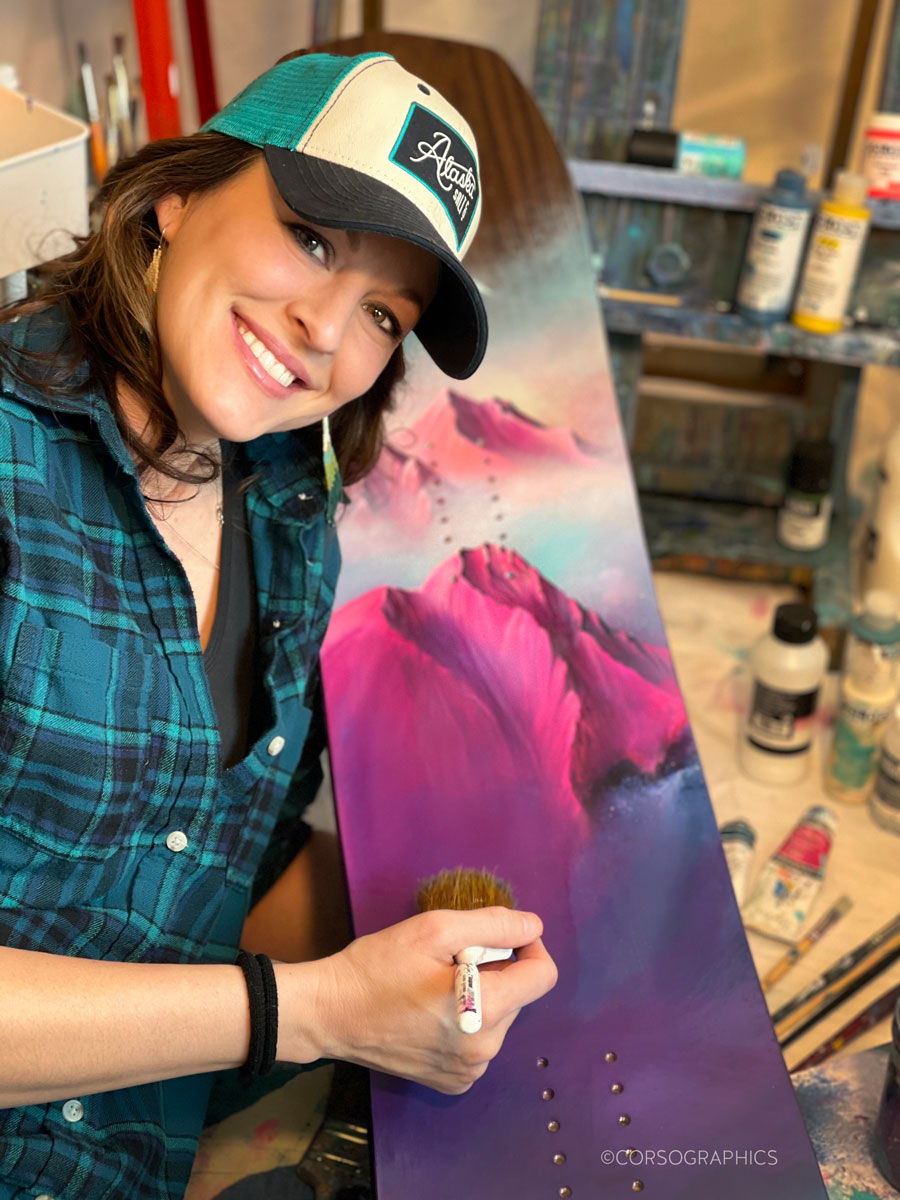 Woman painting a colorful mountain scene on a snowboard.