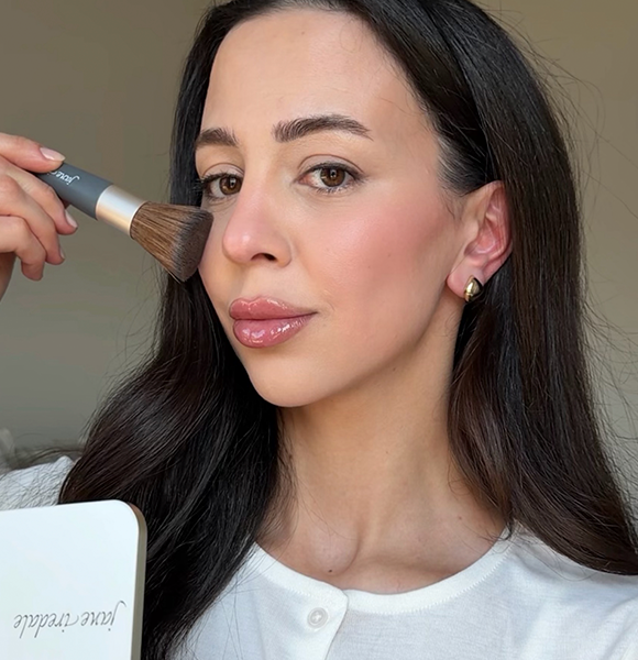 Woman applying makeup with a brush, looking at the camera.