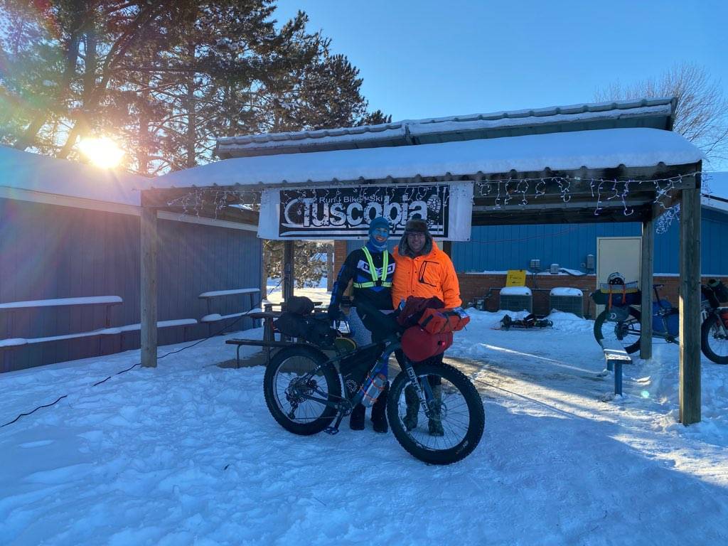A smiling cyclist and her husband stand behind her fully-loaded bikepacking bike at the finish line of the Tuscobia 160.