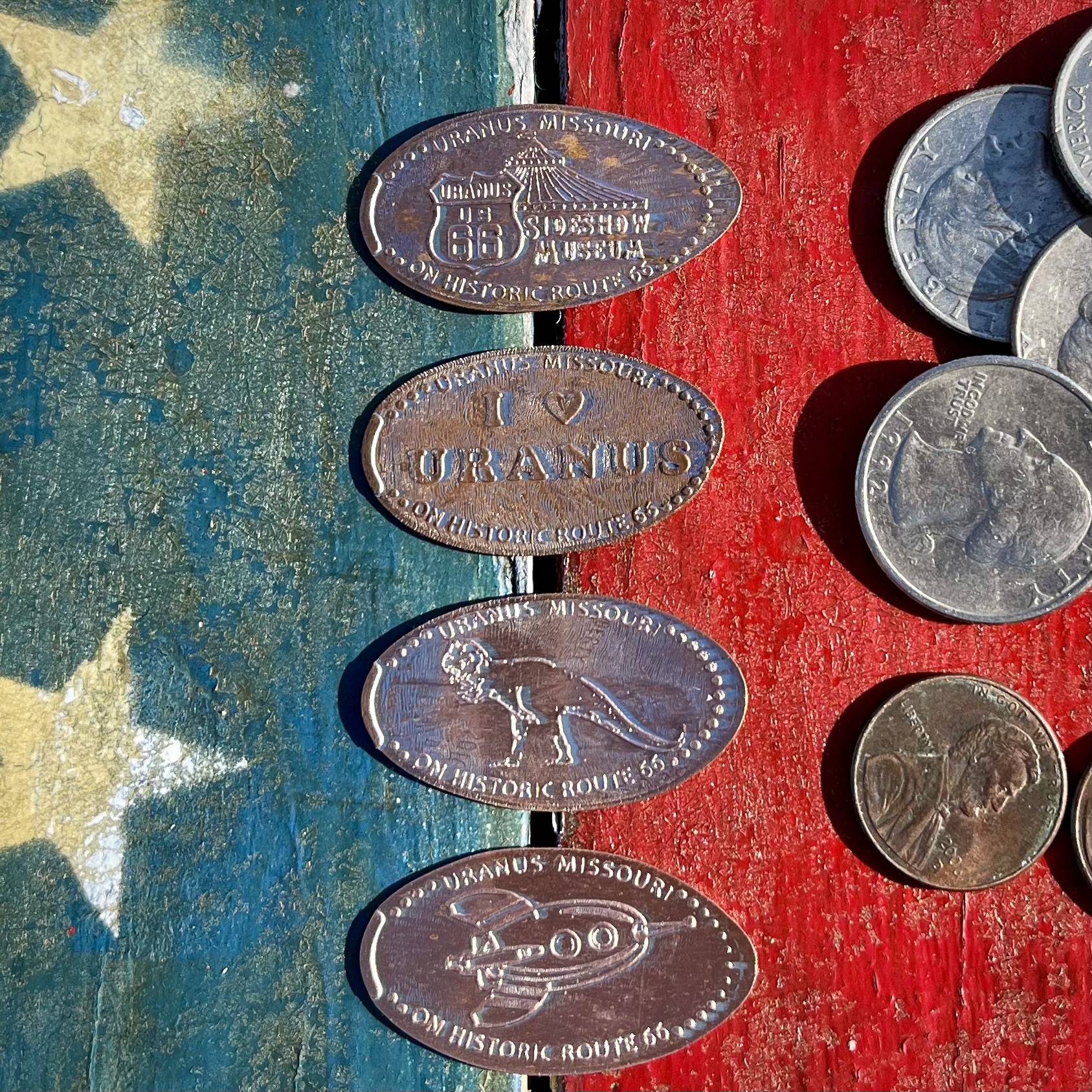 Four unique pressed pennies from Uranus Fudge Factory on Route 66 in Missouri lie on an American flag backdrop, along with two pennies and a few quarters. 