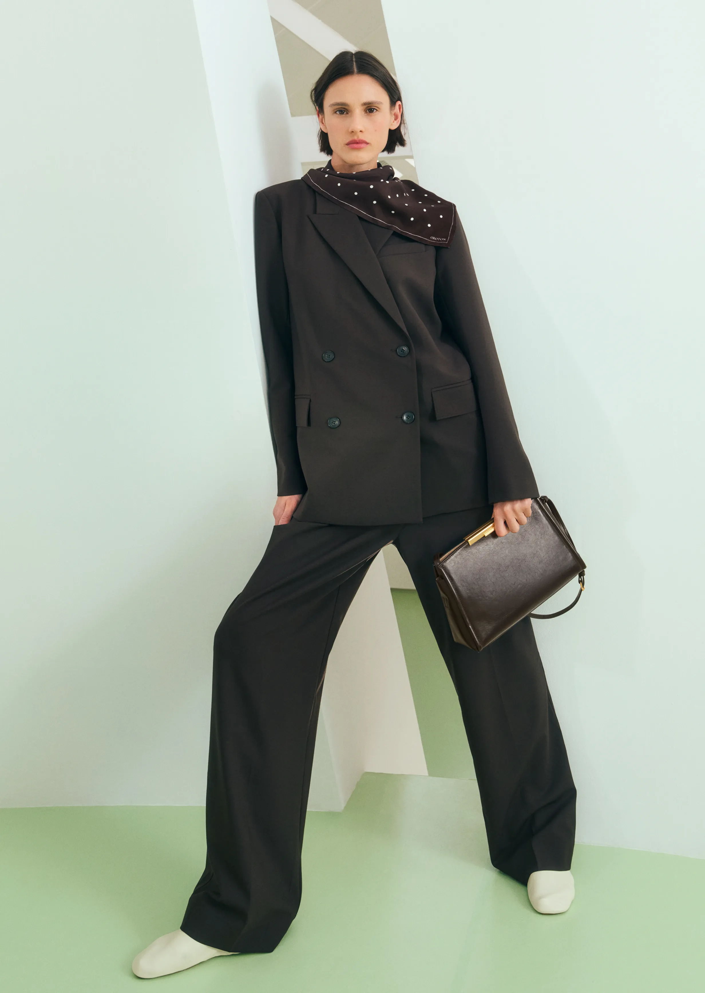 Model in a black suit holding a handbag, standing against a geometric background.