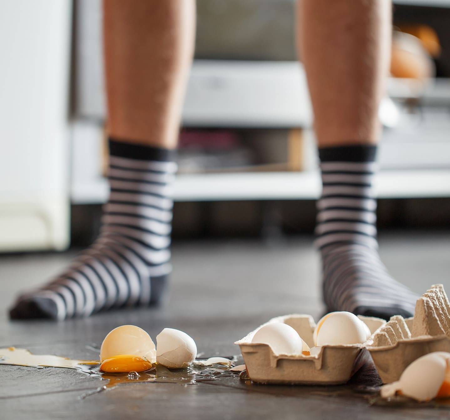 Man in stripey socks stands over a box of cracked eggs. He may or may not experience egg allergy symptoms 