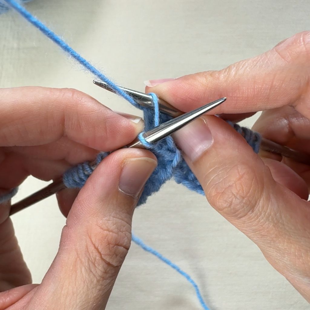 Detailfoto, wie eine rechte Masche gestrickt wird. Mit der rechten Nadel wird der Faden durch die Masche geholt.