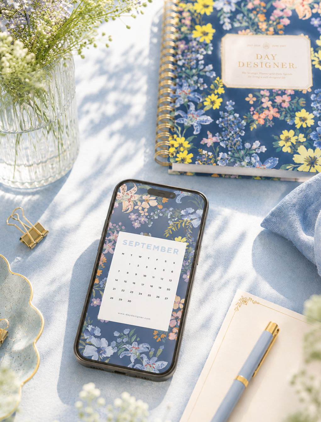 september 2026 digital wallpaper shown on phone, blue table with floral planner, vase with flowers, dish, notepa with blue pen