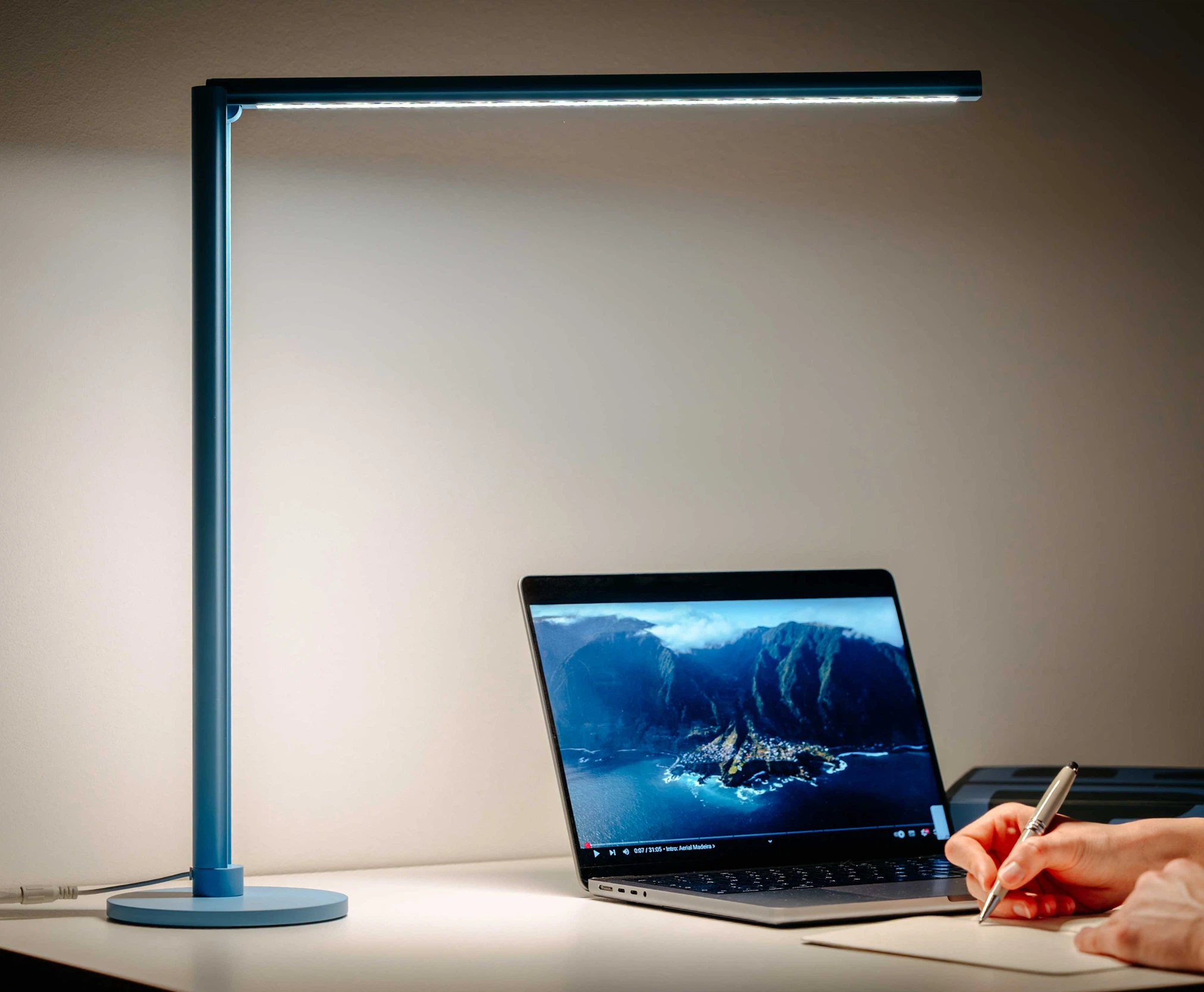 Lumino 's blue desk lamp illuminates a workspace with an open laptop and a handwritten note on a white table.