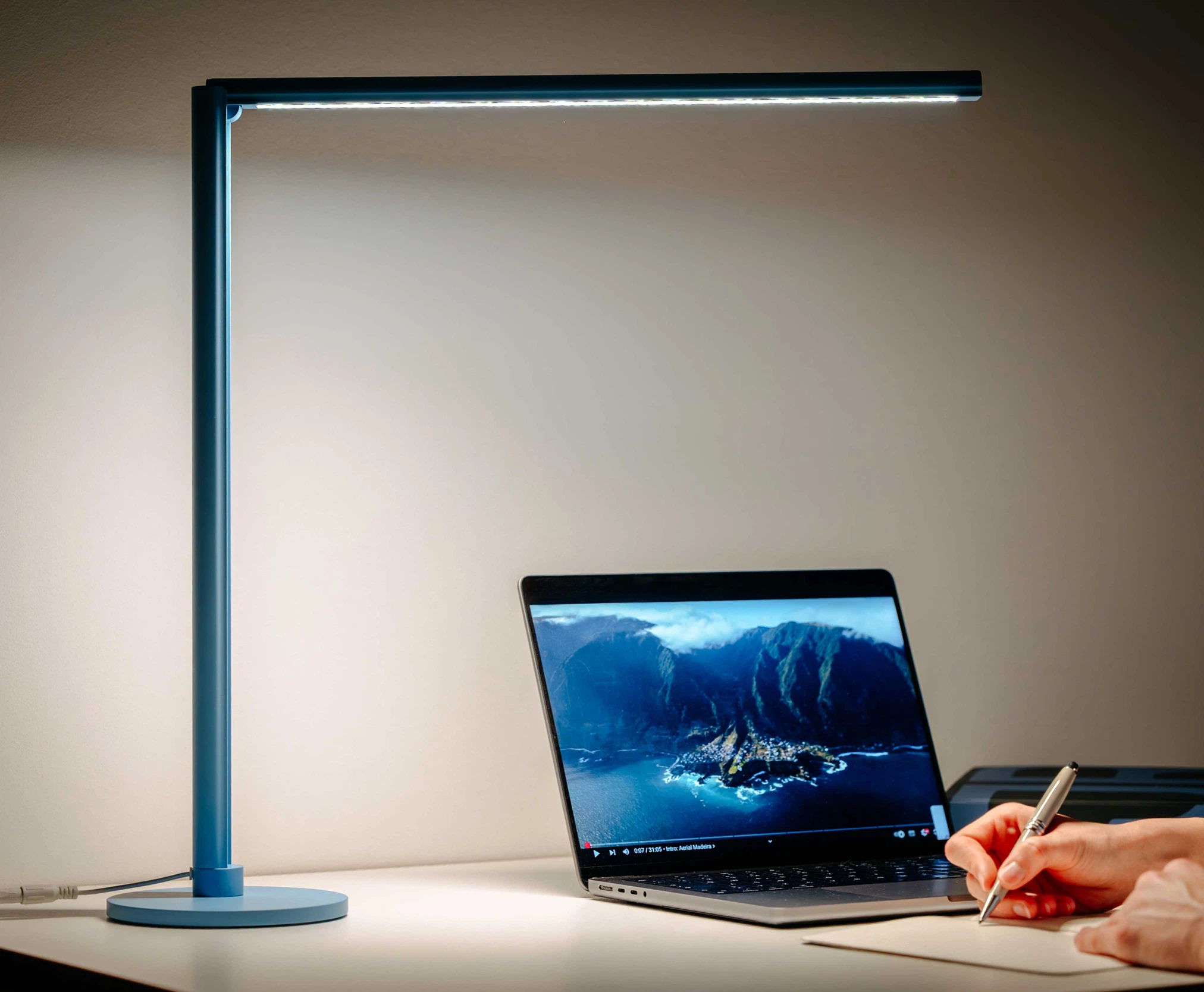 Blaue Schreibtischlampe Lumino Desk beleuchtet einen Arbeitsplatz mit geöffnetem Laptop und einer handgeführten Notiz auf einem weissen Tisch.