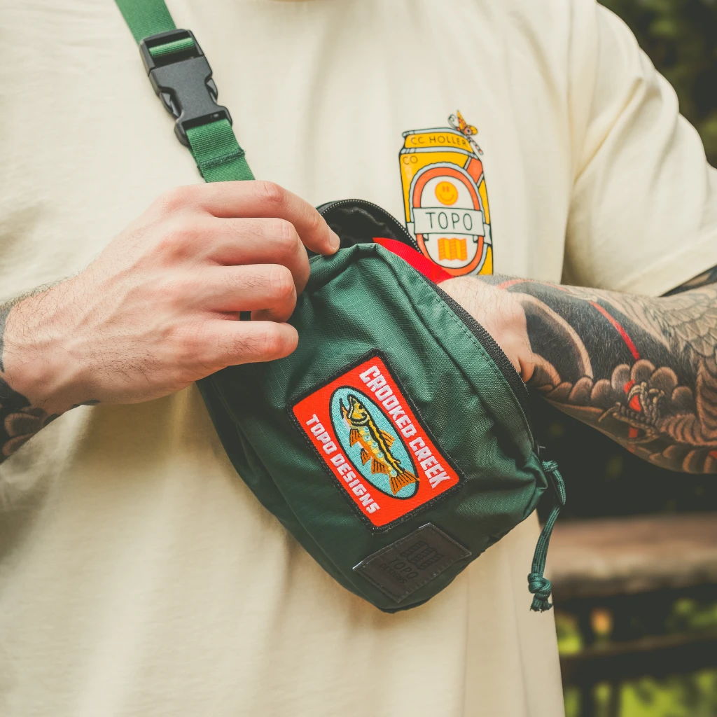Person wearing a tan shirt, holding a green bag with a patch.