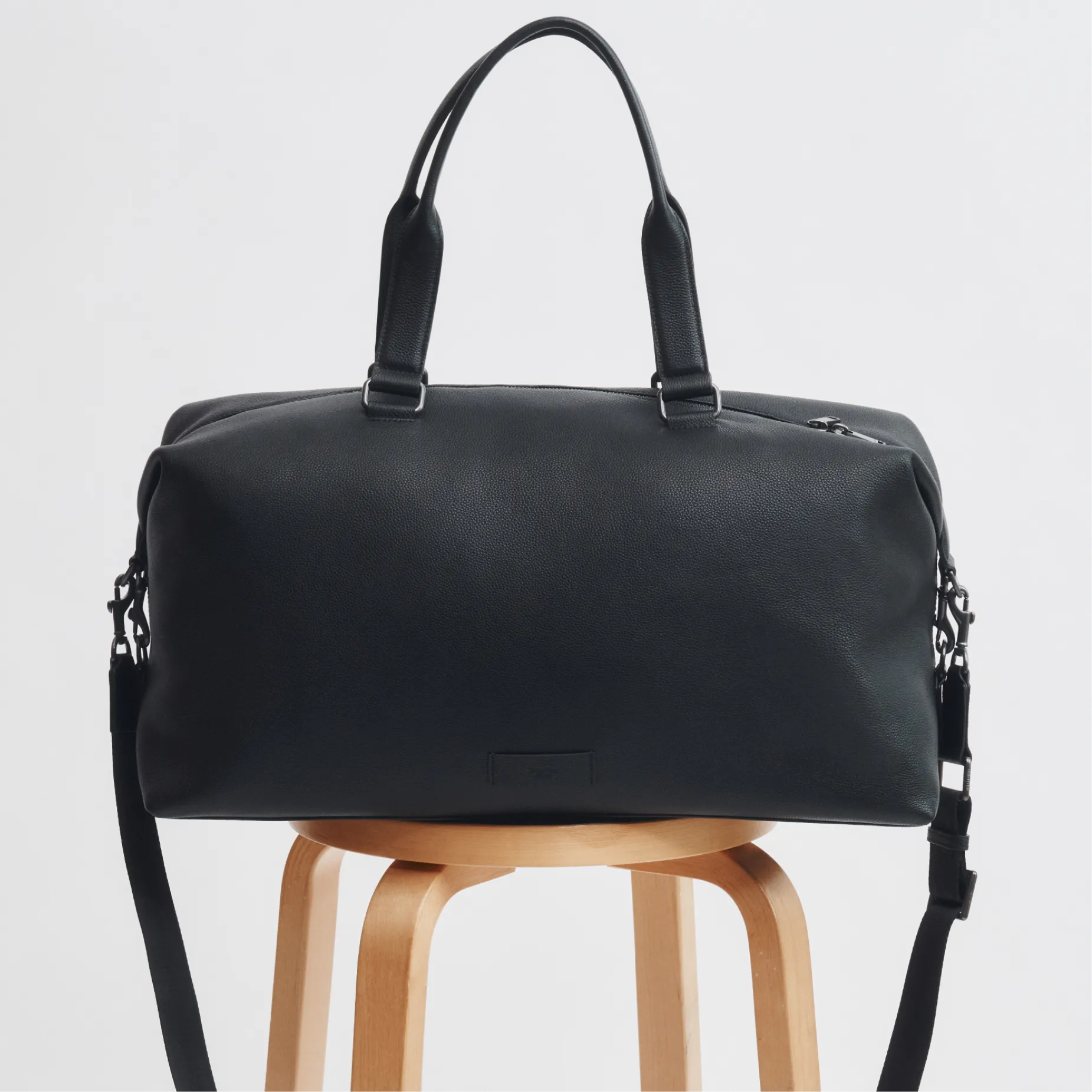 Black leather duffel bag on a wooden stool against a white background.