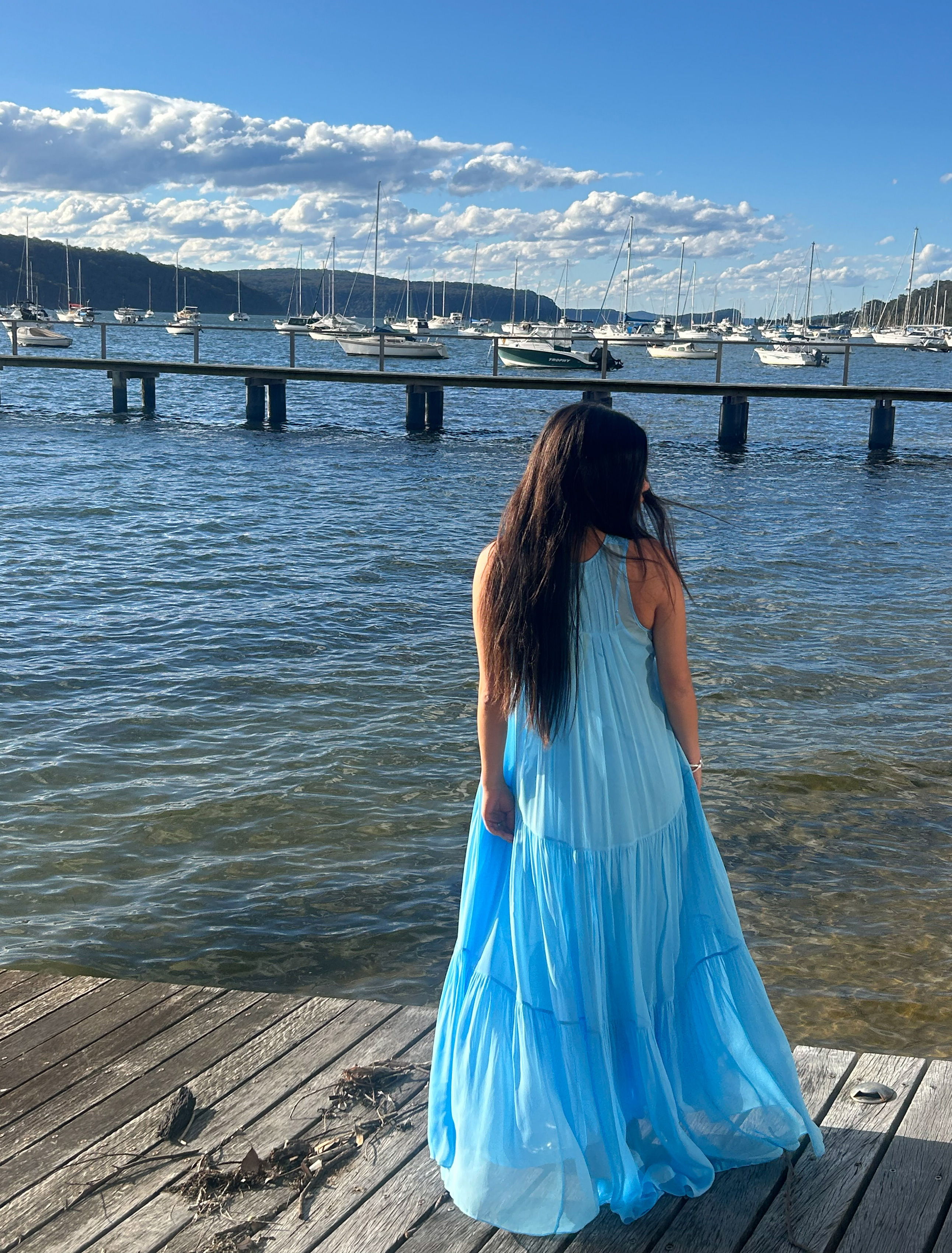 Person in a blue dress standing by the water, looking at boats.