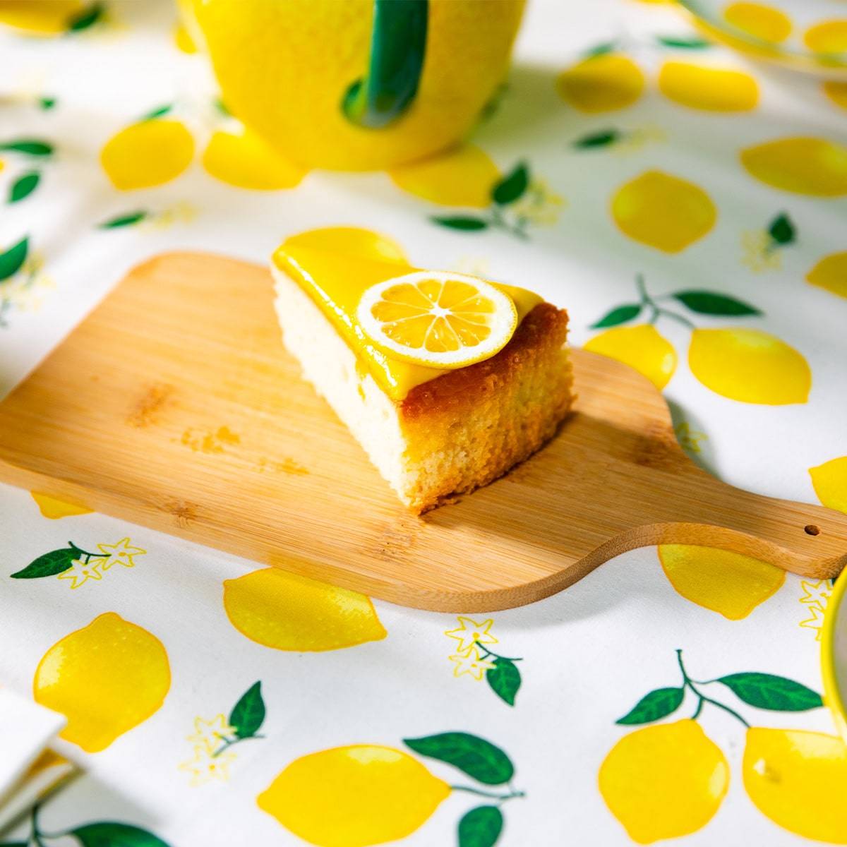 A delectable slice of lemon cake topped with a thin lemon slice, resting on a bamboo cutting board. The cake and board are set against a vibrant background featuring a lemon-print tablecloth, complementing the theme with scattered lemon illustrations and green leaves, creating a fresh and appealing summer dessert setup.