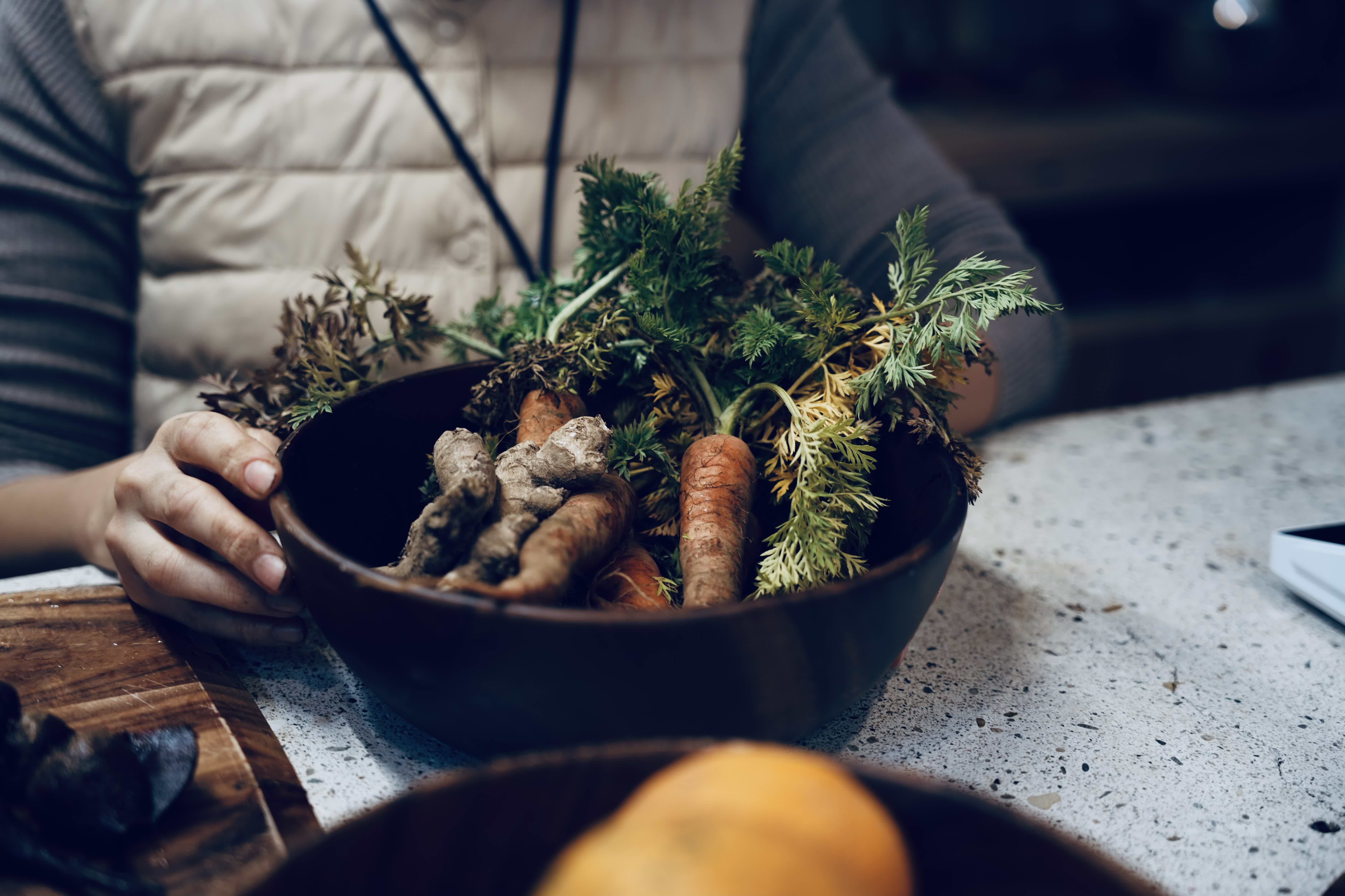 Person holding a bowl of fresh root vegetables and greens on a table.