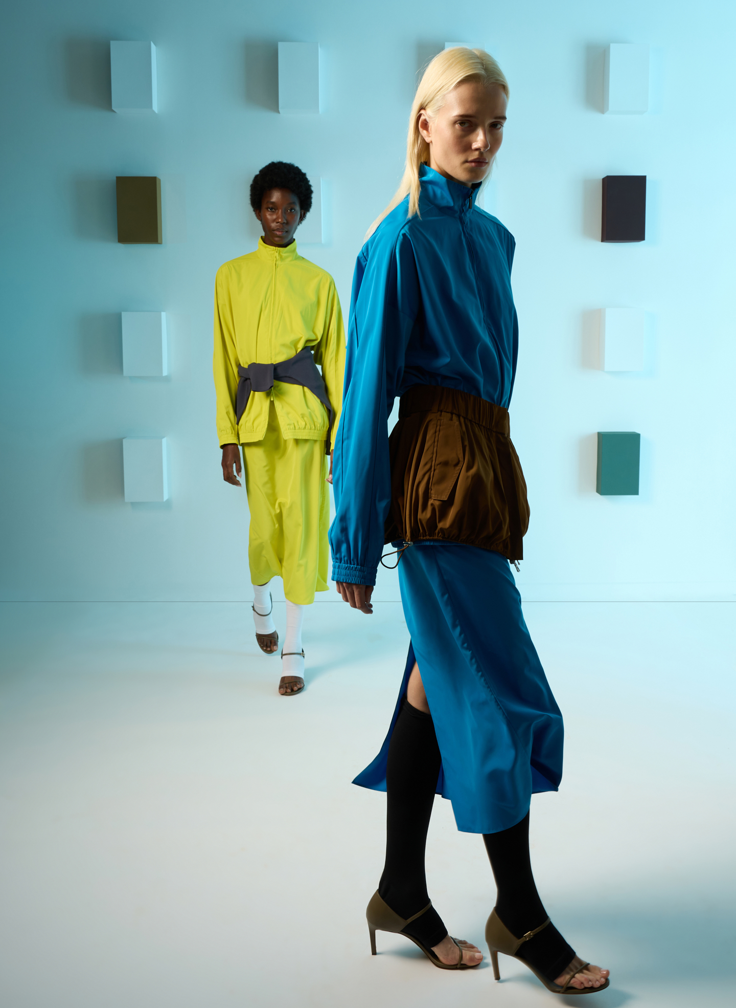 Two models in colorful outfits pose in a minimalist setting.