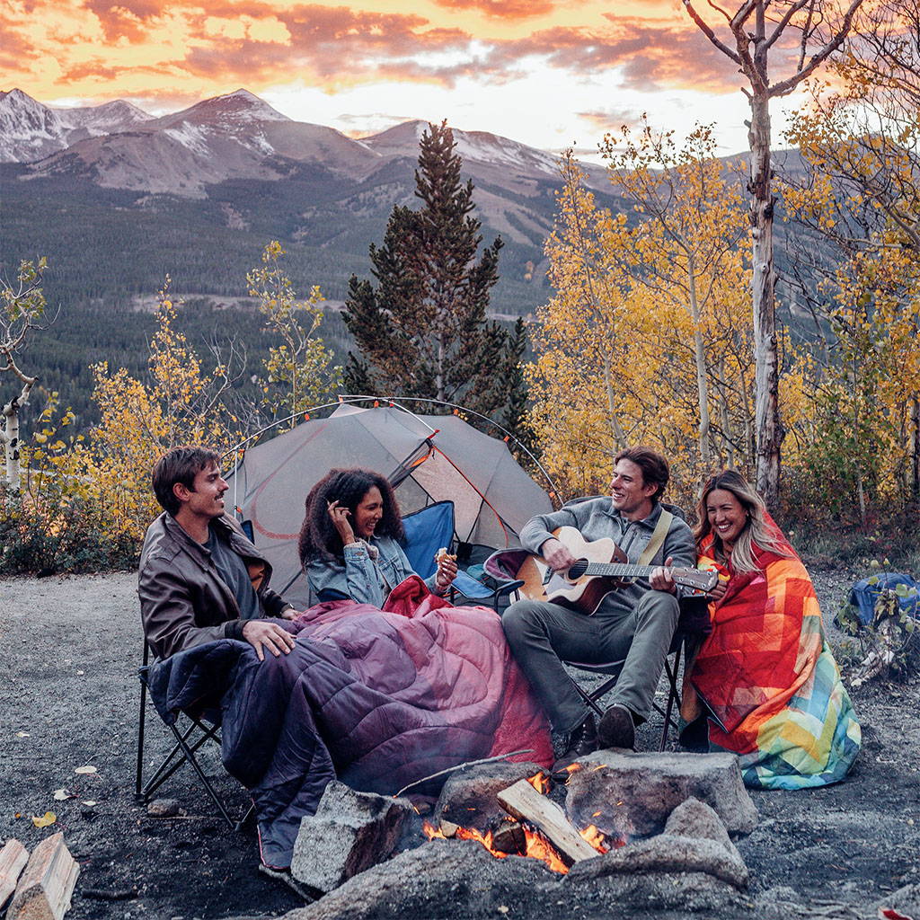 Four friends sit by a campfire, sharing Rumpl blankets to keep warm in the outdoors.