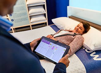 Back of man holding computerised tablet seen in foreground. Smiling woman lies on SleepPRO bed.