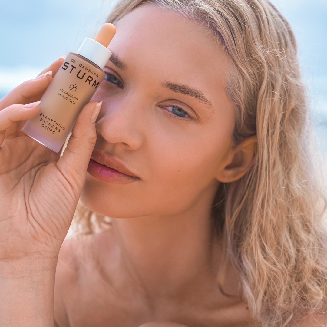 A woman holding a foundation bottle near her face at the beach.