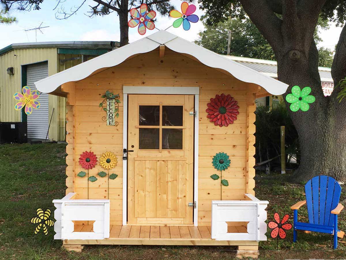 Wooden Playhouse with a white roof and railing and decorative flowers with a blue chair next to it by WholeWoodPlayhouses