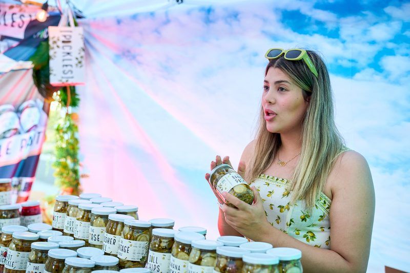 Vendor holding a jar of banana peppers at a Pickle Row booth stacked with hot pickles, kosher dill, dill chips, and vintage pickle jars, part of unlimited pickle sampling at The Big Dill® Philadelphia pickle party at Stateside Live! on National Pickle Day