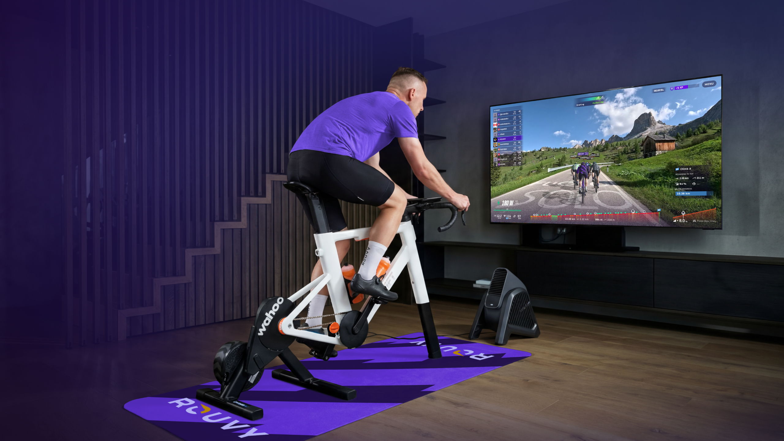 Person trainiert auf einem Indoor-Bike mit Smart-Trainer im Wohnzimmer und fährt mit der Rouvy-App virtuell auf einer Bildschirmanzeige durch eine Berglandschaft.