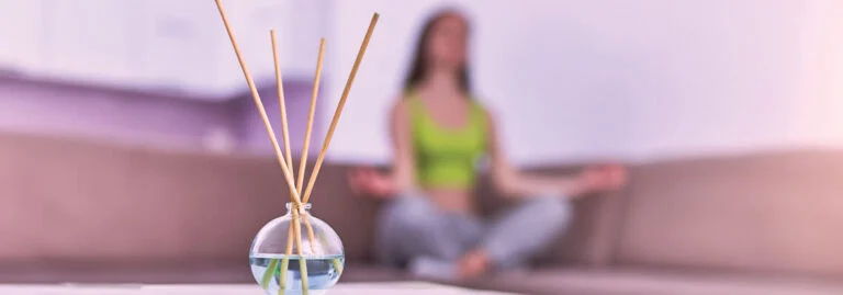 A diffuser with sticks in the foreground and a person meditating in the background.