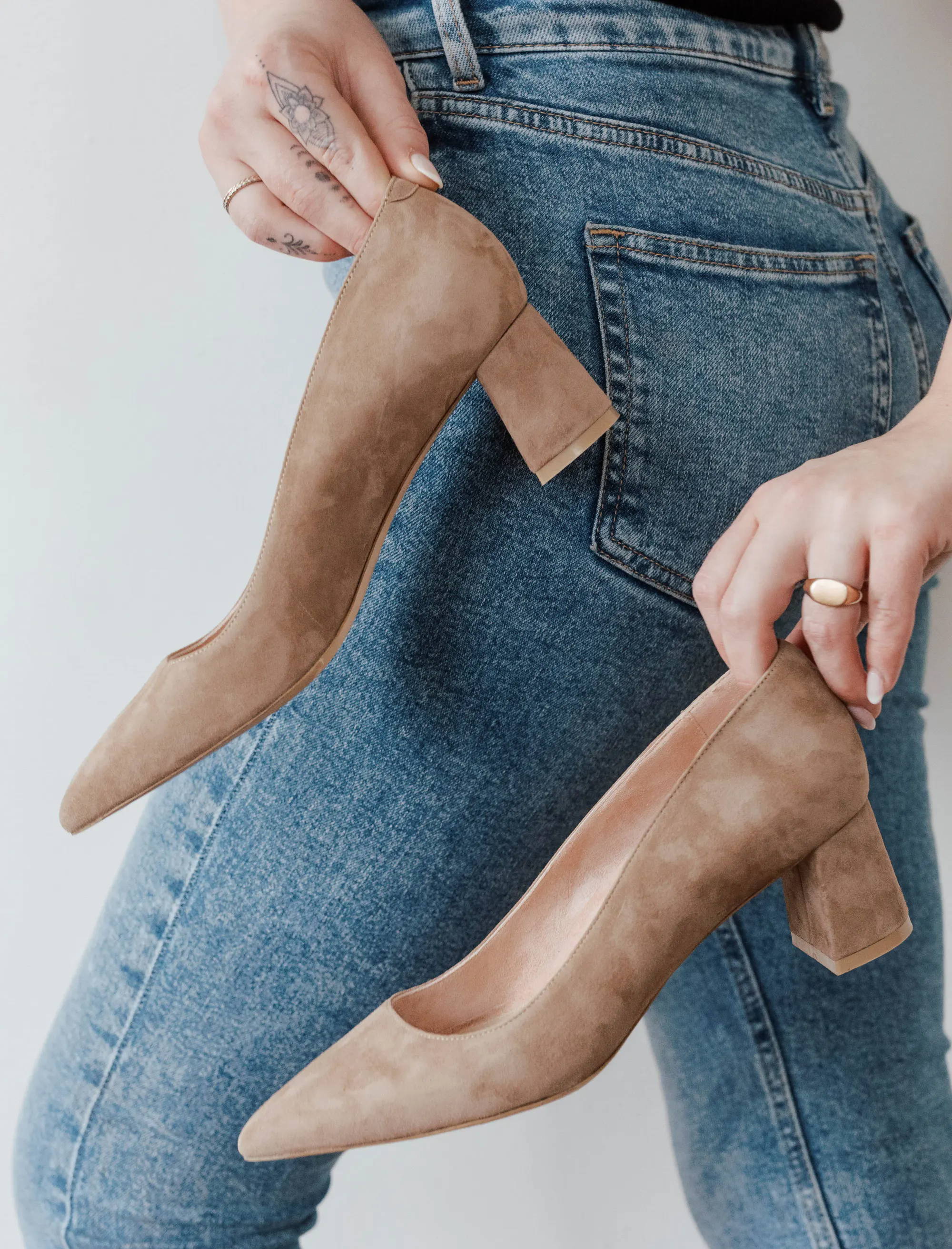 Person holding two pairs of beige high-heeled shoes while wearing blue jeans.