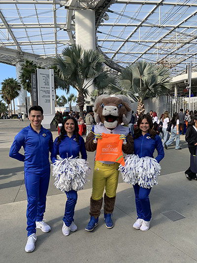 Cheerleaders and a mascot pose in an indoor venue with palm trees.