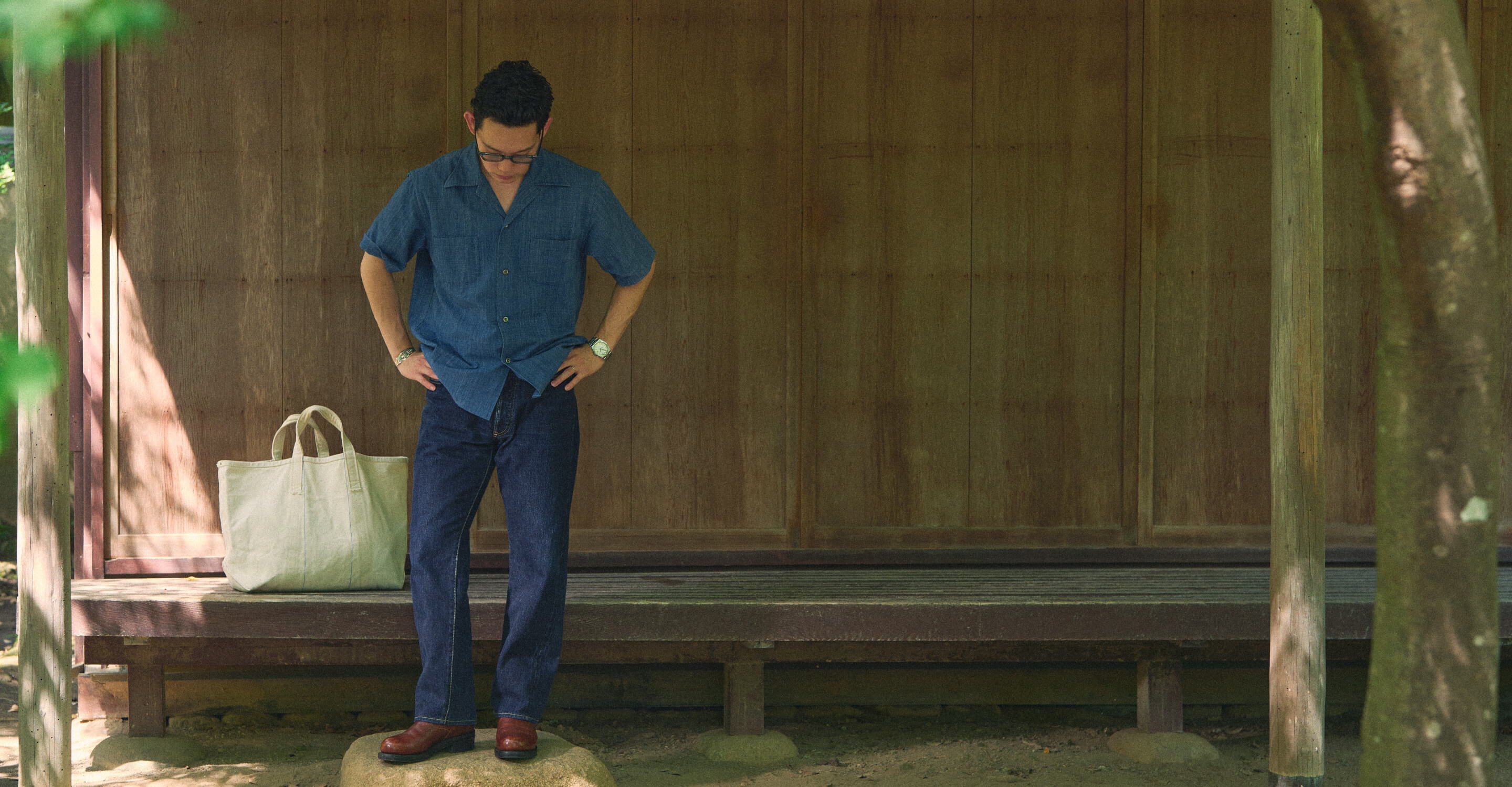 Person standing outside a wooden structure, looking down, with a bag nearby.