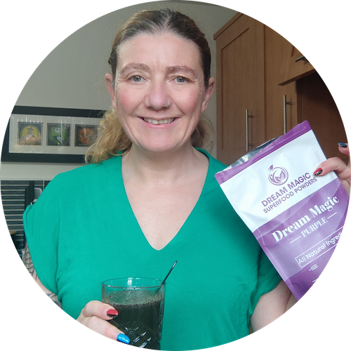 A woman stands in a home kitchen holdinng a pack of Dream Magic Purple blend and a glass of Dream Magic. 