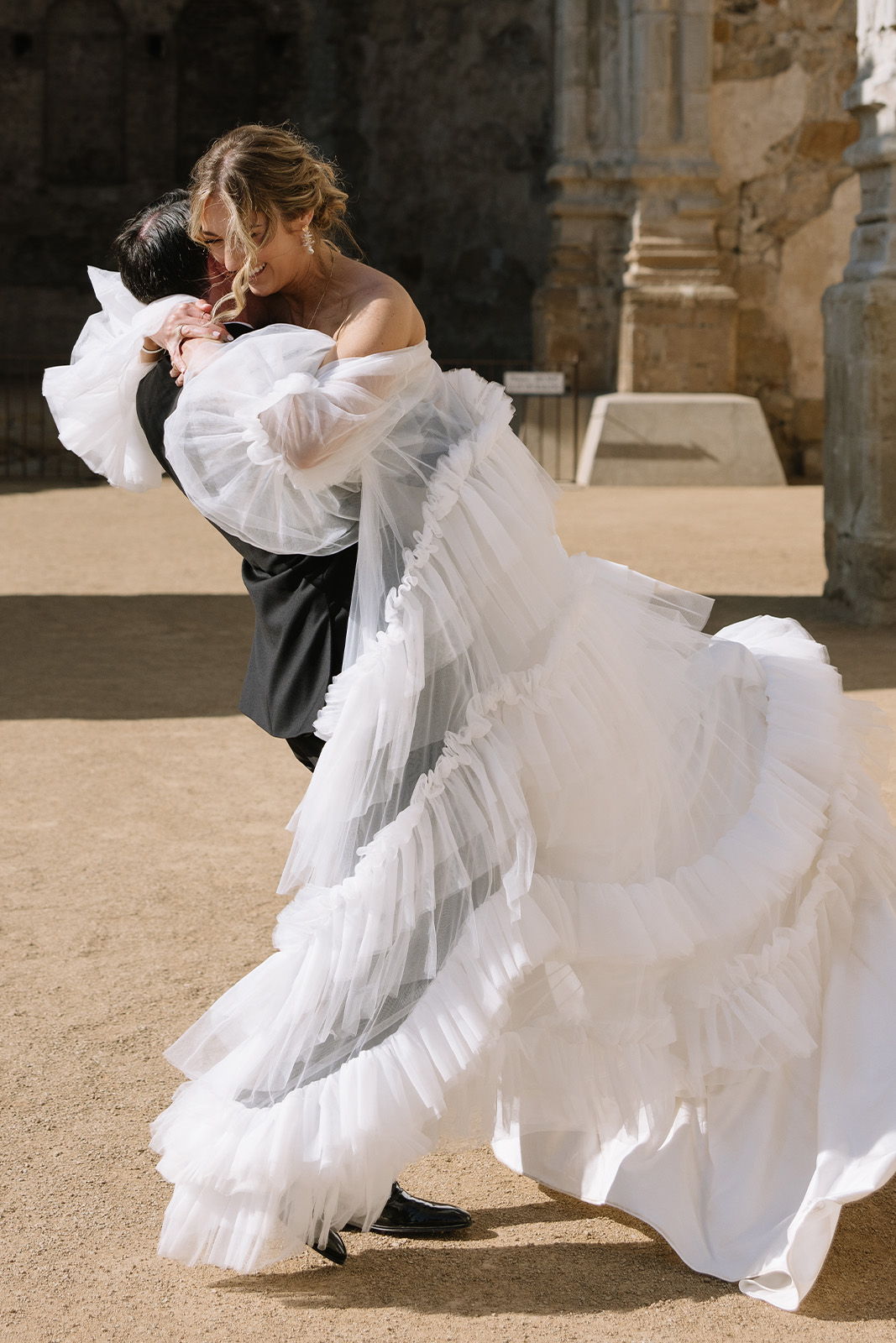 Couple embracing, one in a white ruffled dress, outdoor setting.