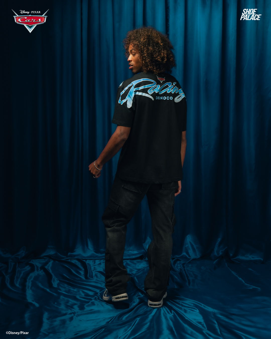 Person with curly hair wearing a black shirt, standing against a blue backdrop.