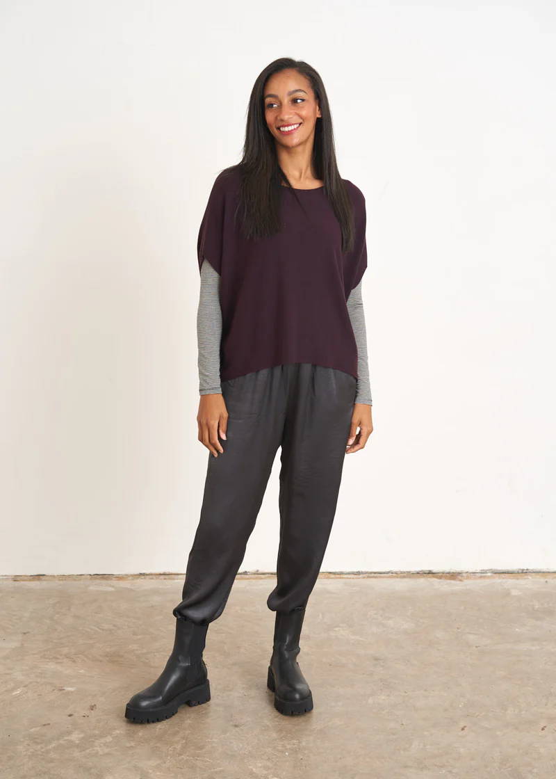 A model wearing a sleeveless, aubergine coloured sweater over a grey long sleeved toip and dark grey satin trousers and black chelsea boots