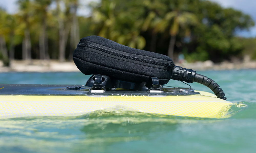 Duotone Battery Box Mounting Set posé sur une planche de surf dans des eaux peu profondes, avec des palmiers en arrière-plan.
