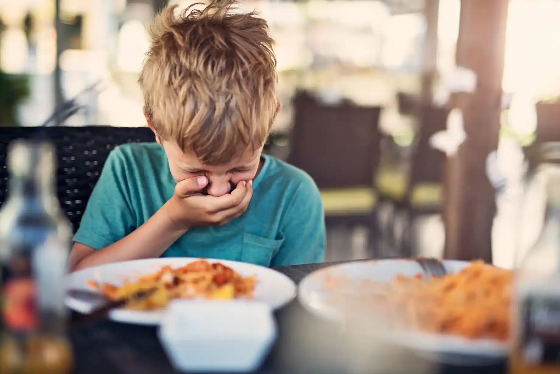 Ein Junge sitzt über seinem Mittagessen und hält sich seinen Mund zu und den Kopf nach unten – Seine Zunge ist angeschwollen und ihm ist übel. Dies könnten erste Anzeichen einer Anaphylaxie sein.