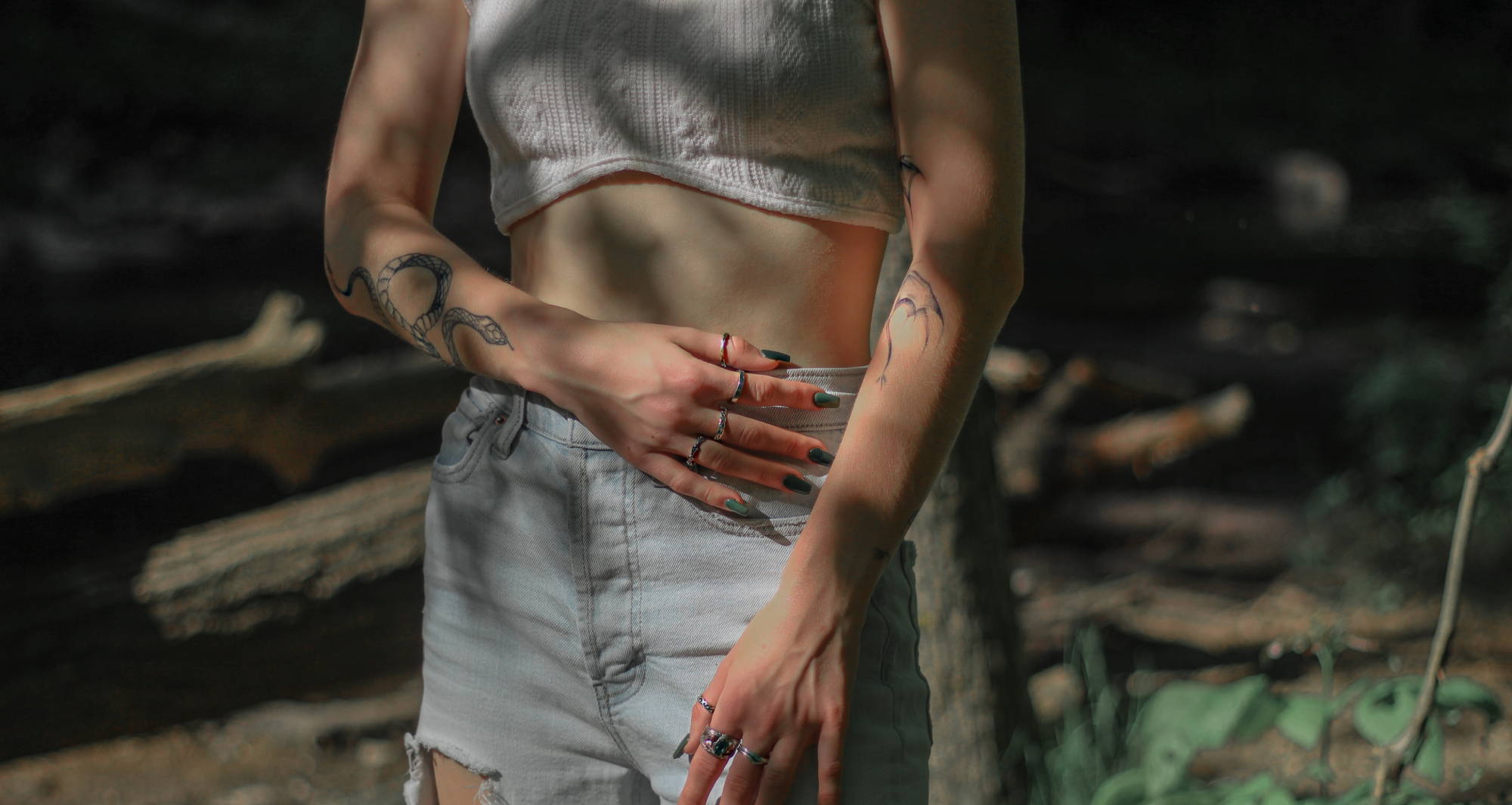 A woman with long, green nails and tattoos, wearing a white crop-top and ripped jeans touches her stomach as she stands in a forest.