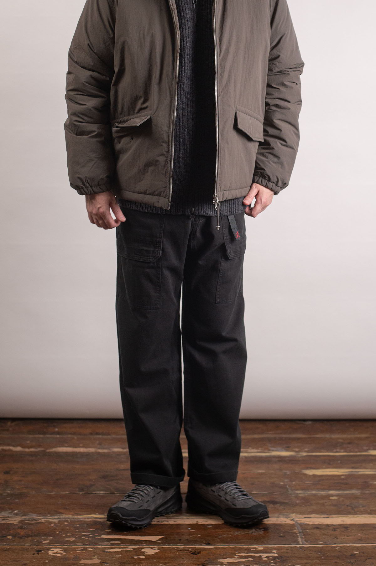 Person wearing a puffer jacket, sweater, black pants, and shoes on wooden floor.