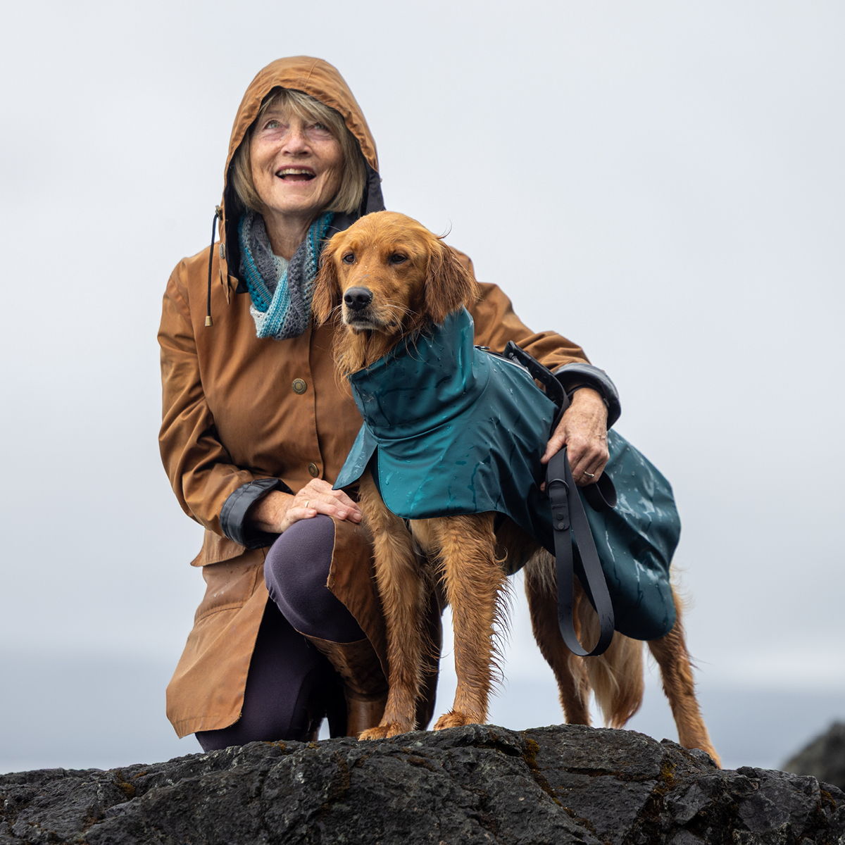 Woman in a raincoat smiling with a dog on a rocky surface.