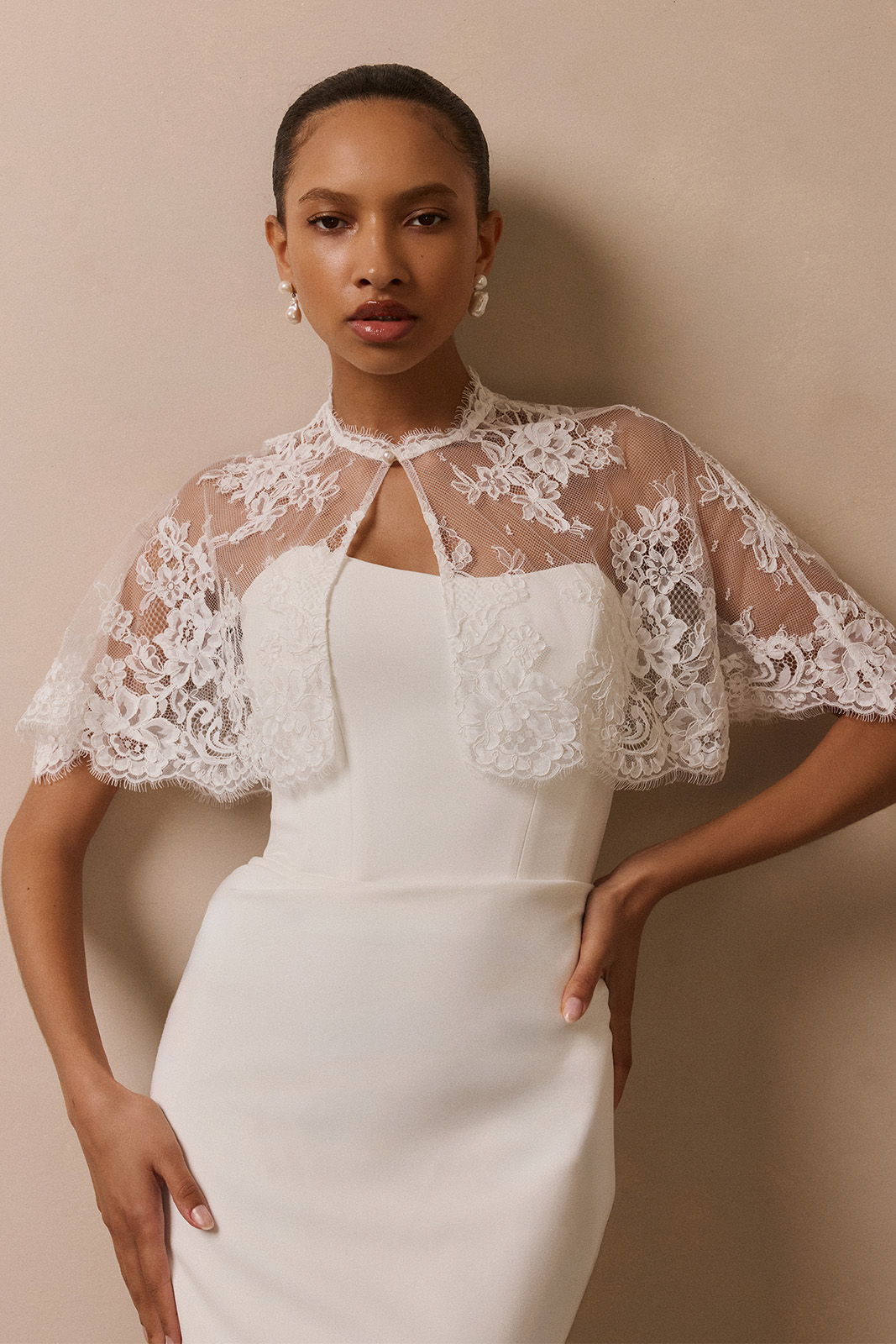 Model wearing a white dress with a lace capelet against a neutral background.