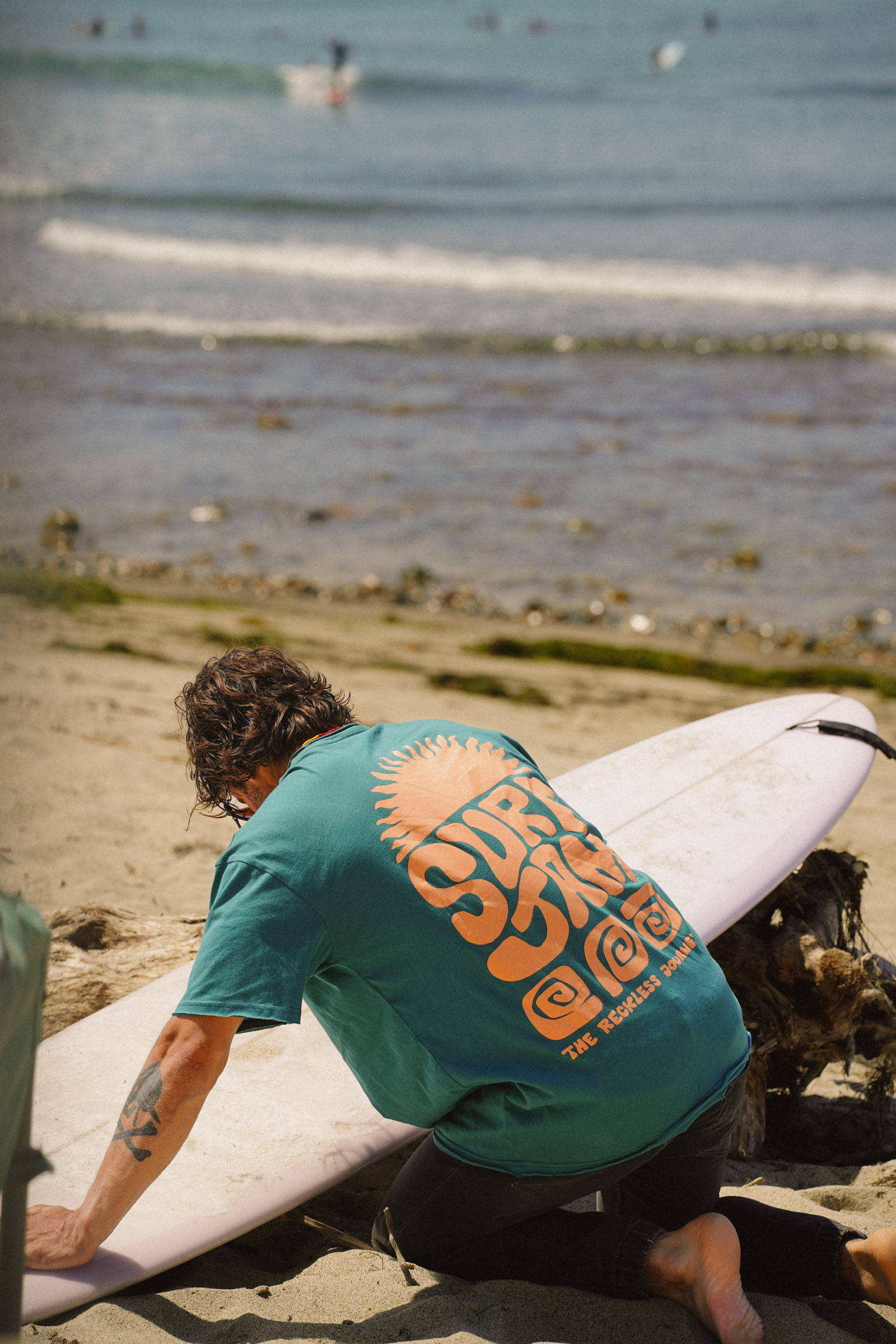 A man waxing his surfboard wearing a cool graphic t-shirt by the ocean.