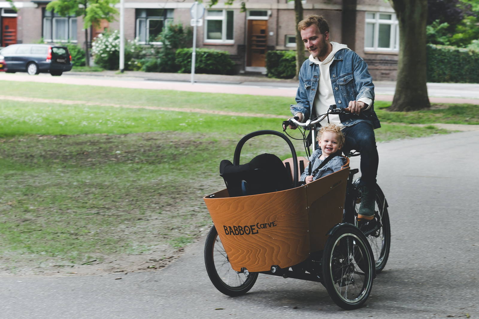 A father rides a Babboe Carve cargo bike with his two young children in the front box. The infant is riding in a Maxi Cosi car seat strapped in with a Maxi Cosi Adapter.
