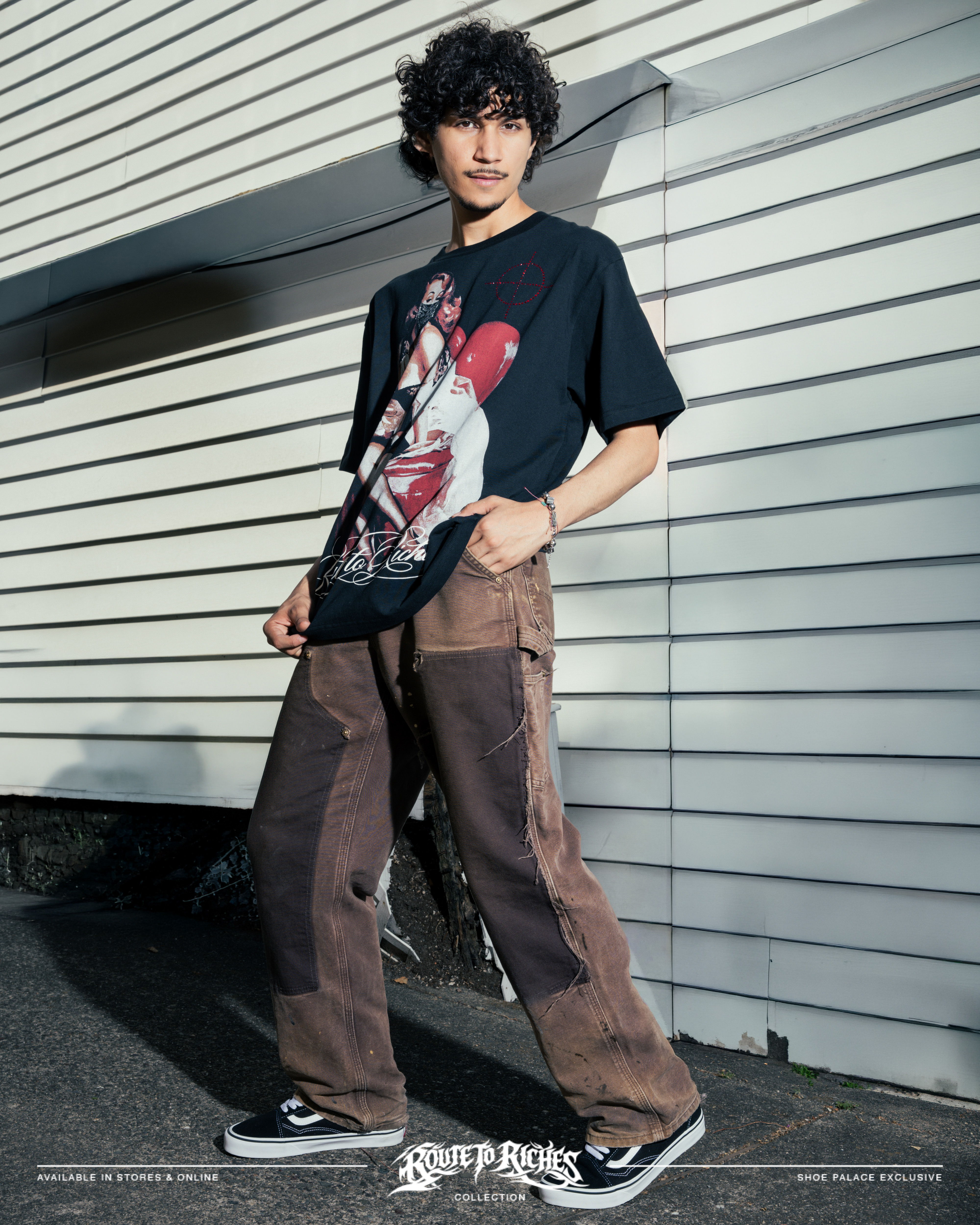 A person wearing a graphic t-shirt and baggy pants stands against a wall.