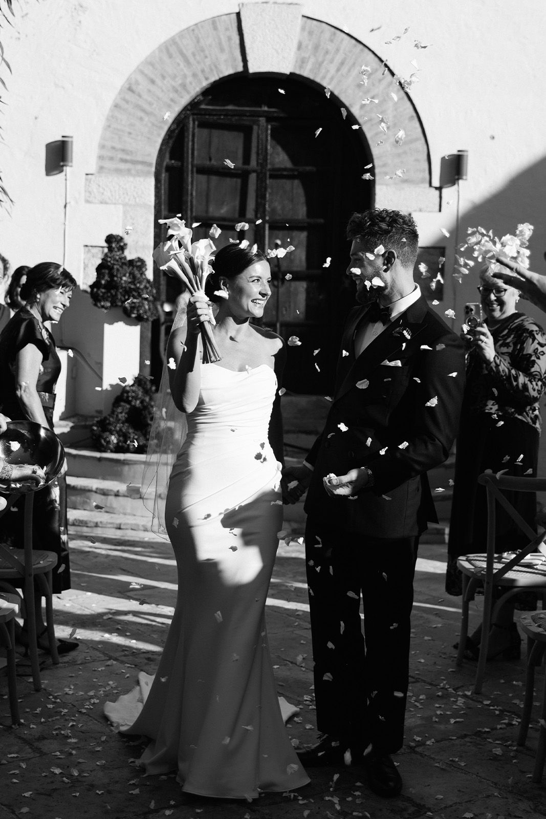 Bride and groom celebrating with confetti outside a venue. Black and white photo.