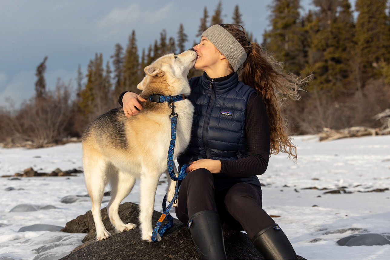 Woman sitting with dog in snowy landscape, sharing a joyful moment.
