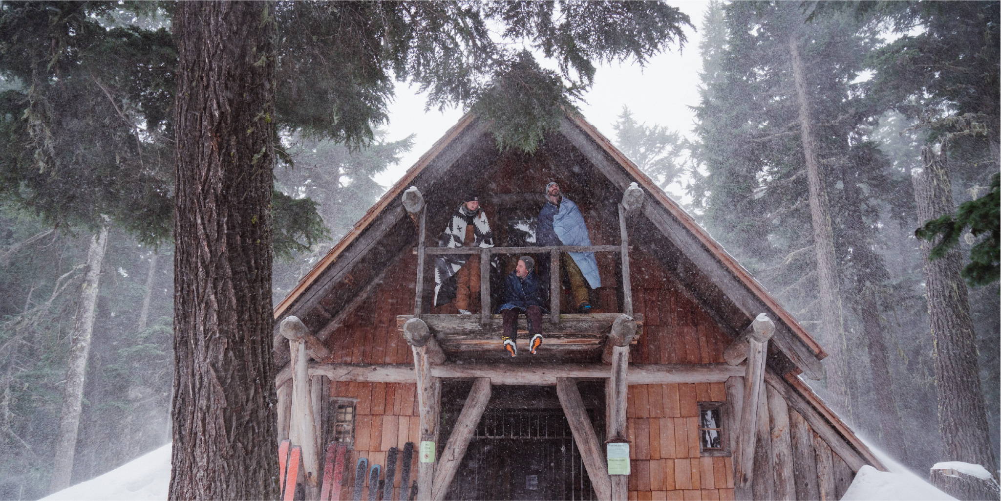 Three people sitting on snowy balcony outside of A frame cabin with snow blanket